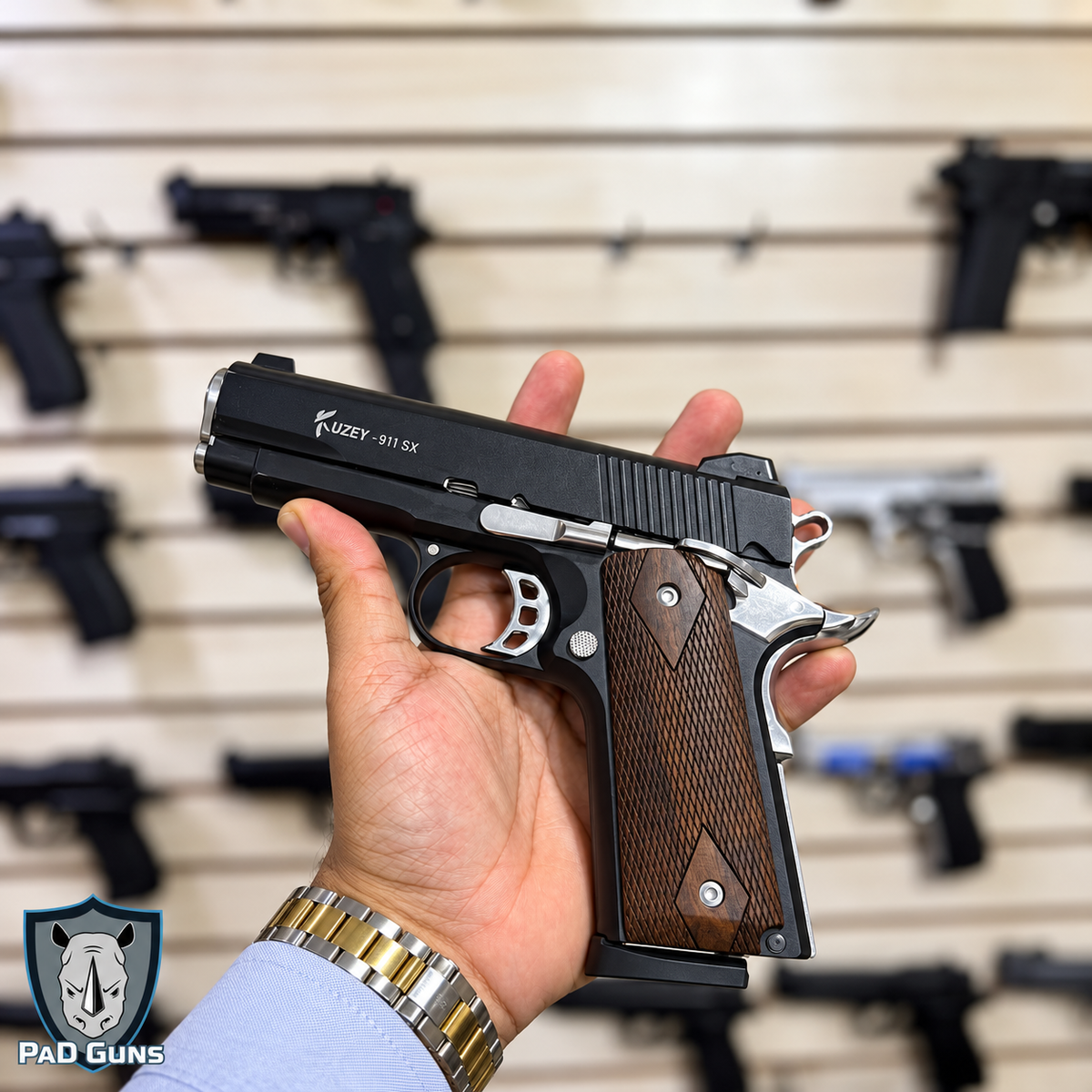 Pistola traumática Kuzey 911 SX bitono en mano, corredera negra con detalles plateados cromados y cachas de madera oscura cuadriculada, estilo clásico 1911, en tienda PaD Guns